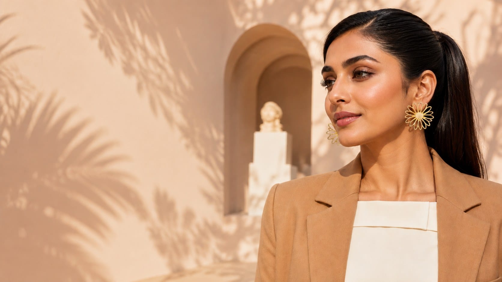 Model in a camel blazer and gold daisy earrings, glancing toward a sunlit archway